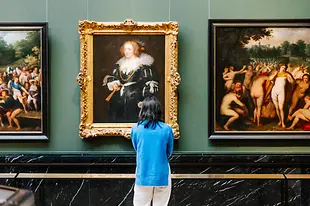 Woman with dark hair a blue shirt and white pants looks at a historic artwork in a golden frame at the Art History Museum in Vienna
